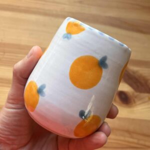 Håndlaget keramikkrus med hvit glasur og håndmalte appelsiner i gult med blågrønne blader, laget av Caroline Sagafos.